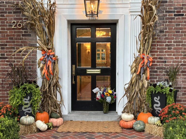 Front door framed by white trim and brick, with harvest decor including cornstalks, pumpkins, and mums on the steps.