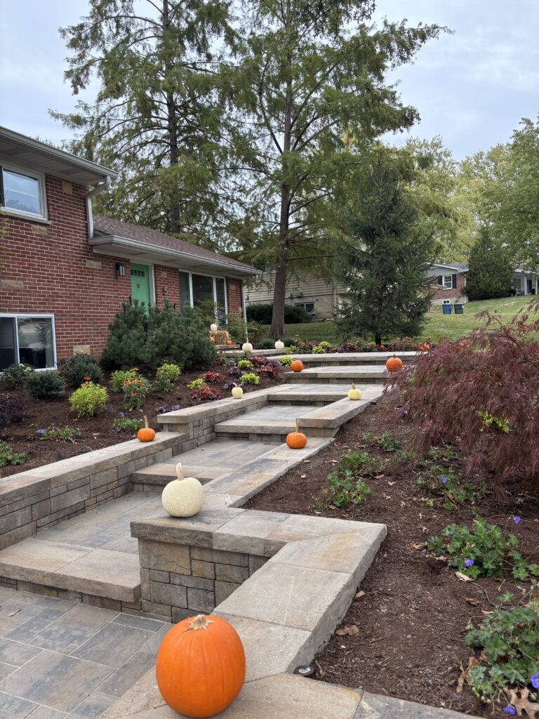 Autumn pumpkins line a multi-level stone staircase in a suburban front yard with a brick house and landscaped flower beds.