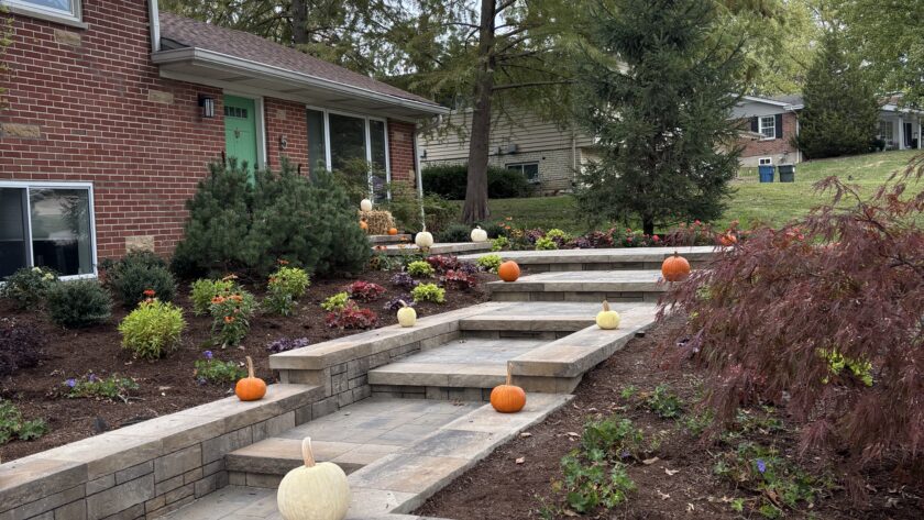 Autumn pumpkins line a multi-level stone staircase in a suburban front yard with a brick house and landscaped flower beds.