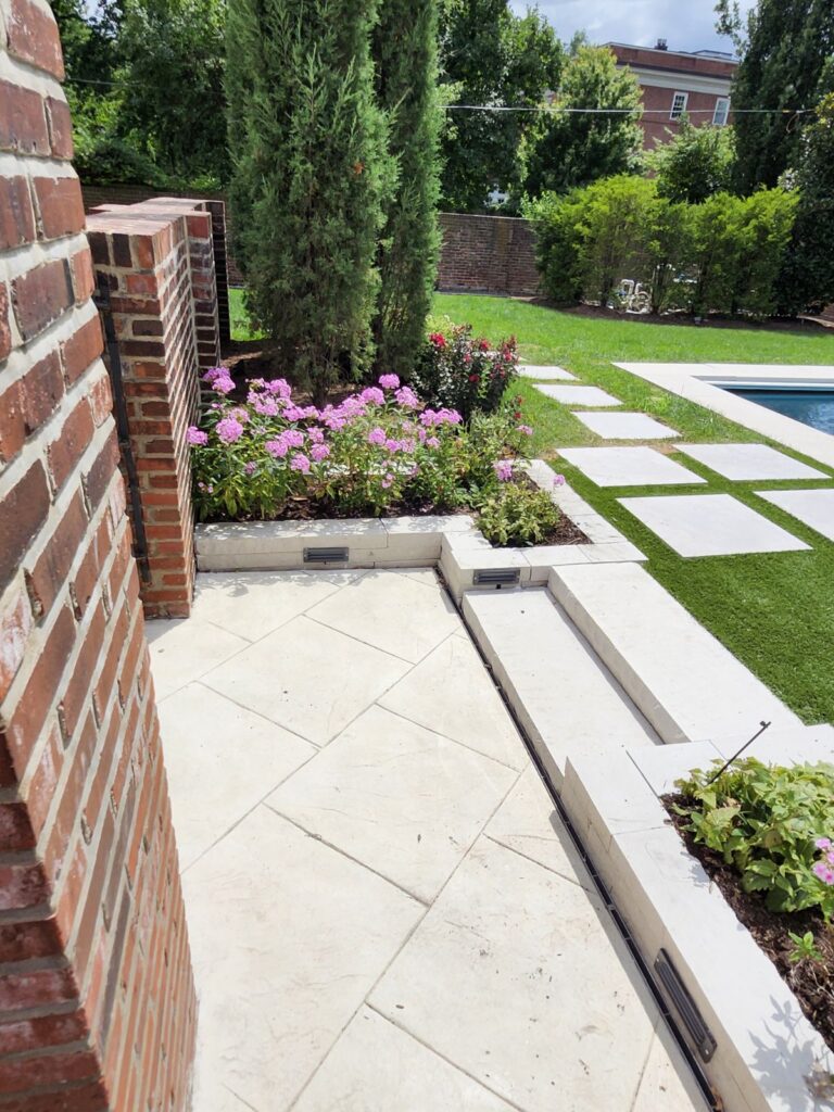 Brick wall and light stone patio with pink flowering bushes, tall evergreens, and a rectangular pool with stepping-stone path across the lawn.