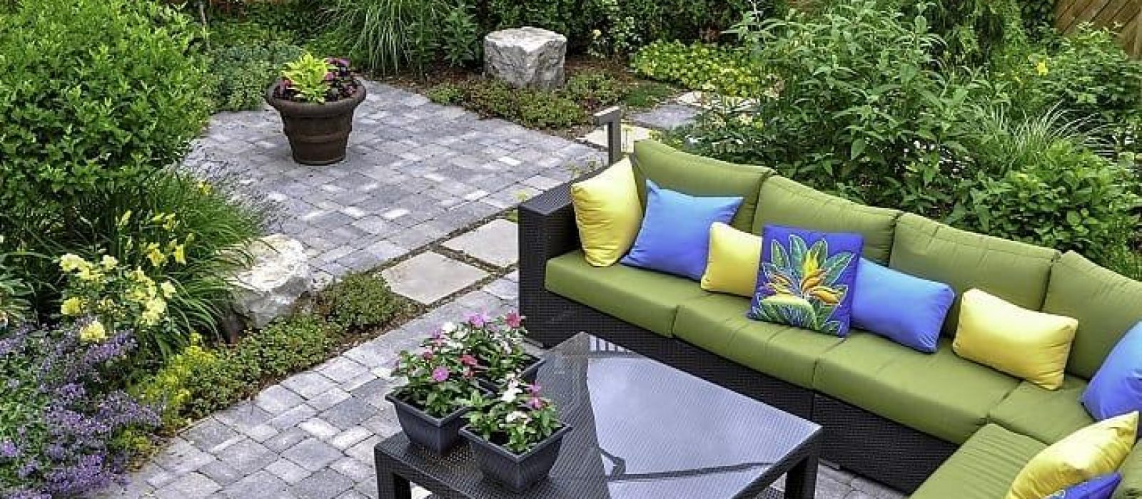 Outdoor patio with green cushioned wicker sectional, colorful pillows, a glass coffee table, and a surrounding garden.